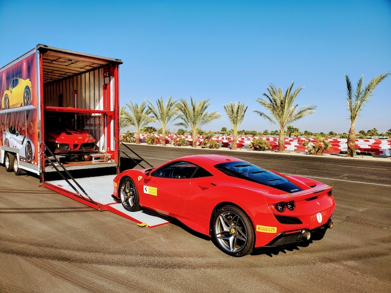 Red sports car being unloaded from a transport truck on a sunny day with palm trees in the background.