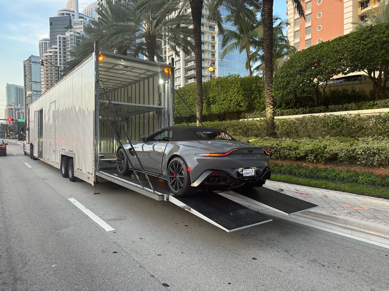 A sports car exiting an enclosed trailer on a city street with palm trees and buildings in the background.