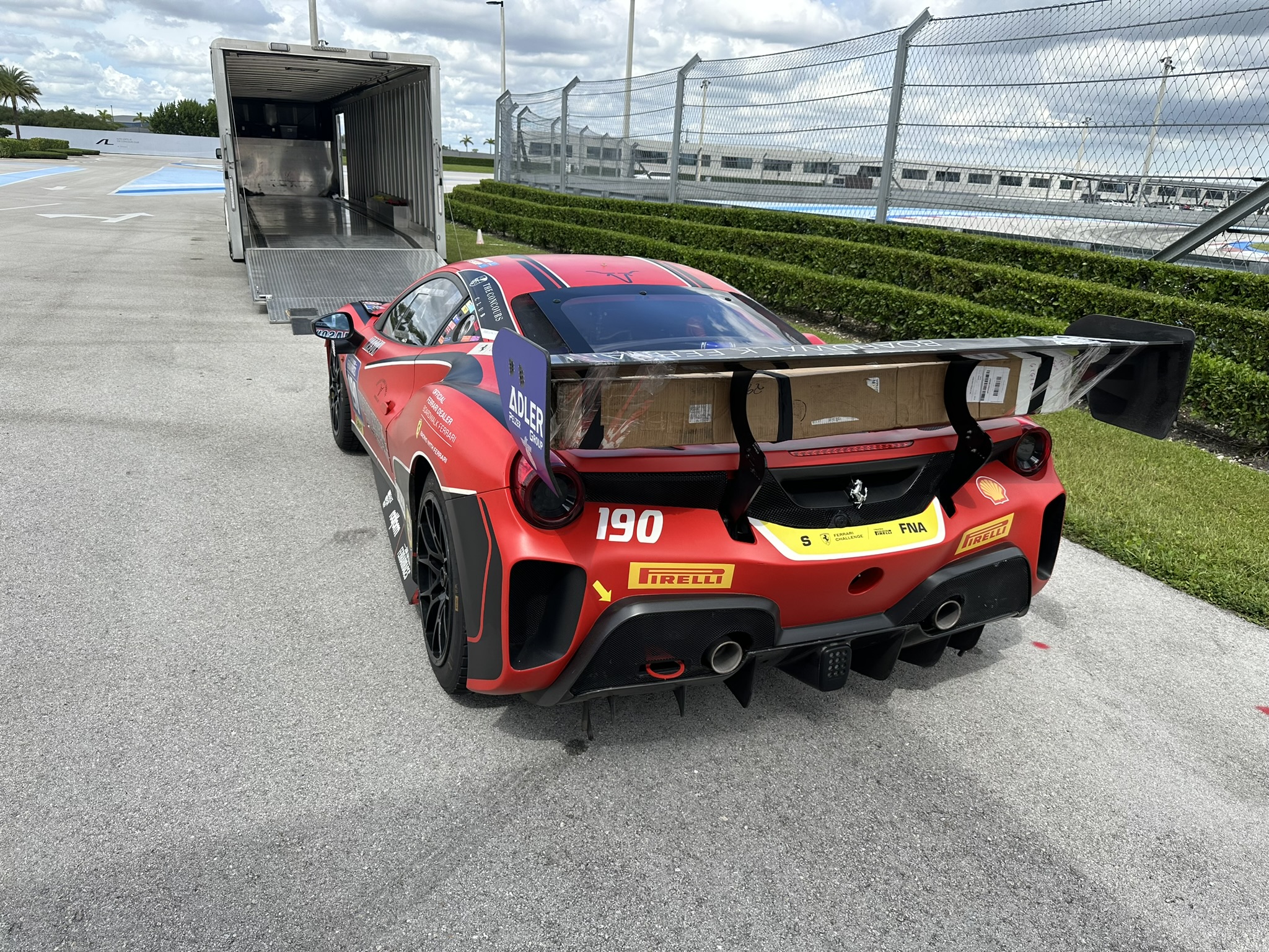 Red Ferrari race car with sponsor decals parked near a transport truck on a sunny day.