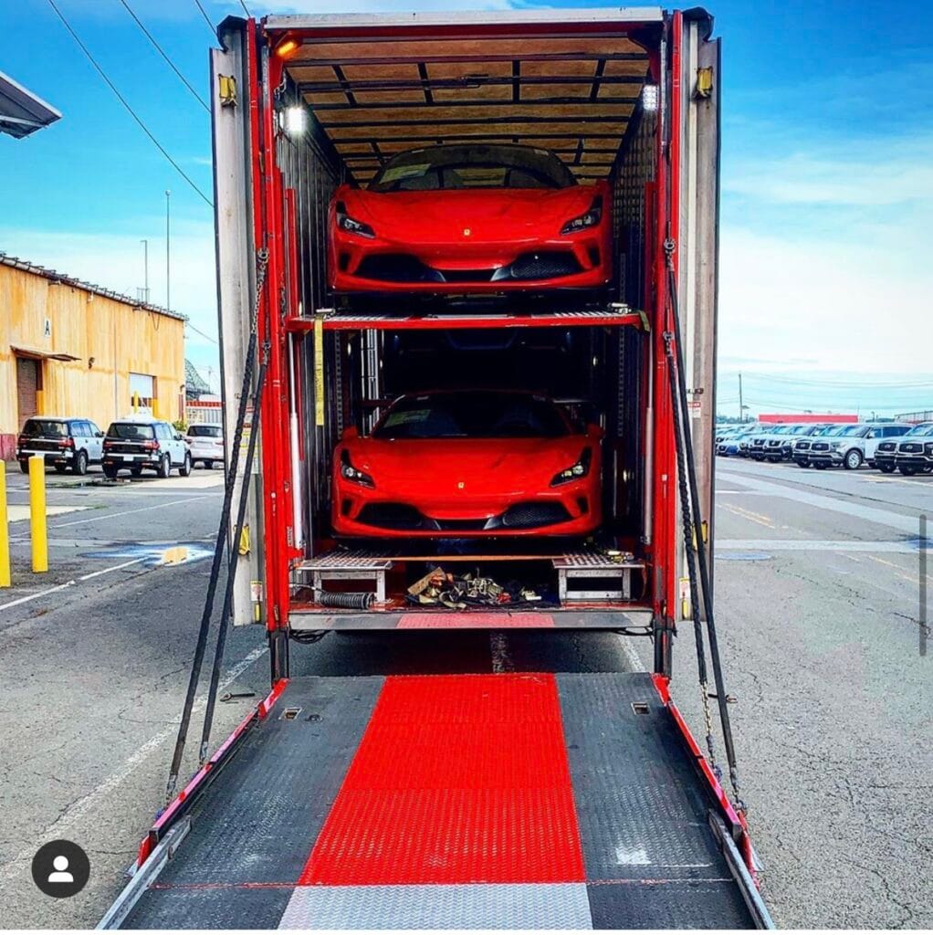 "Open auto transport trailer carrying two red sports cars, parked in an industrial area."