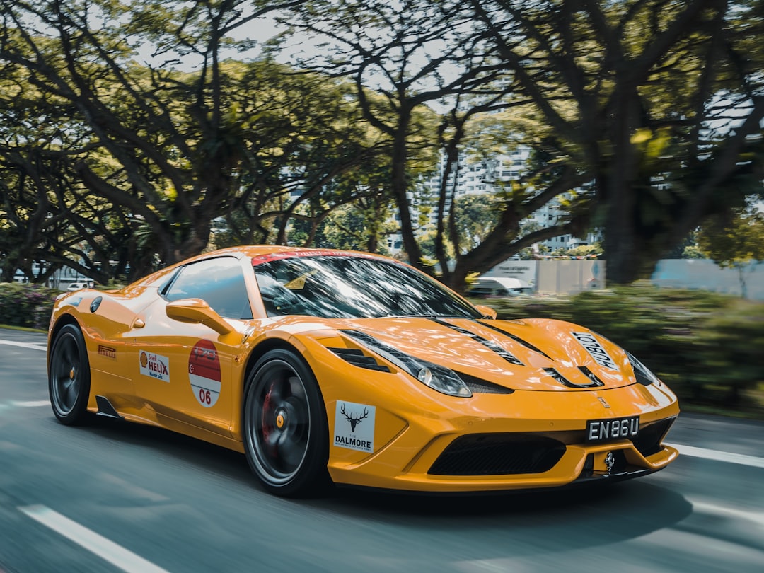 A bright yellow sports car speeds down a shaded road lined with trees, showcasing its racing stickers and visible license plate "EN86U." The background is slightly blurred, capturing the thrill of the ride. Why ship your car with Luxury Auto Transport when you can experience such exhilaration firsthand?