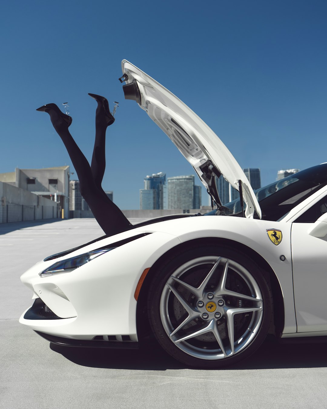 A pair of legs in black stockings and heels humorously protrudes from the open front trunk of a white Ferrari parked on a rooftop. City buildings rise in the background against a clear blue sky, highlighting why it's crucial to choose the best Miami car transport company for luxury cars.