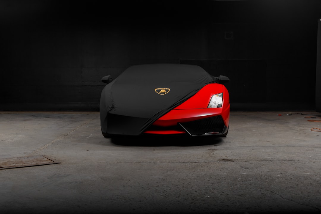 A partially covered red sports car with a visible front bumper and logo is parked in a dimly lit space, hinting at the need for an essential guide to Orlando car shipping. It stands shrouded by a black protective cover against a dark, simple background.