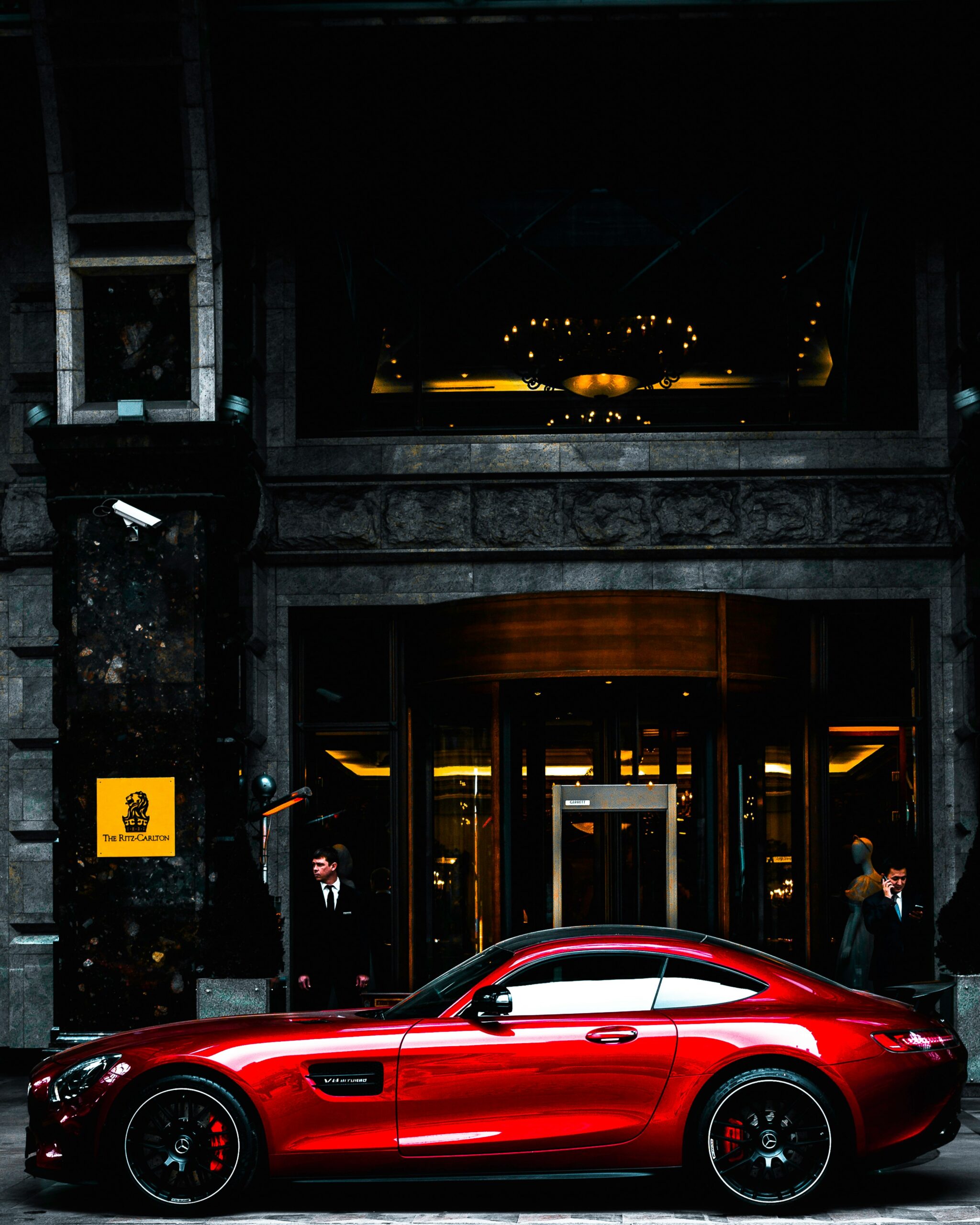 A sleek red sports car is parked in front of a luxurious hotel entrance, showcasing the standard you expect when you choose Luxury Auto Transport. With ornate architecture and large chandeliers inside, two people stand near the entrance as a doorman tends to the scene.