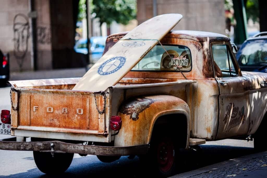 A rusty vintage Ford truck is parked on a street, embodying the spirit of California's surf culture. With a surfboard, logos, and peace sign in its open bed, it offers a glimpse into the state's laid-back vibe― an essential guide to California car shipping through time-worn charm.