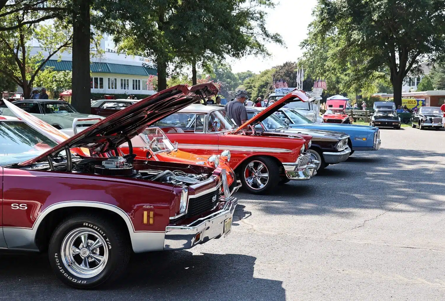 Auto Transport Service Row of beautifully restored classic cars, including a bright red muscle car with its hood open, parked at a sunny outdoor car show.